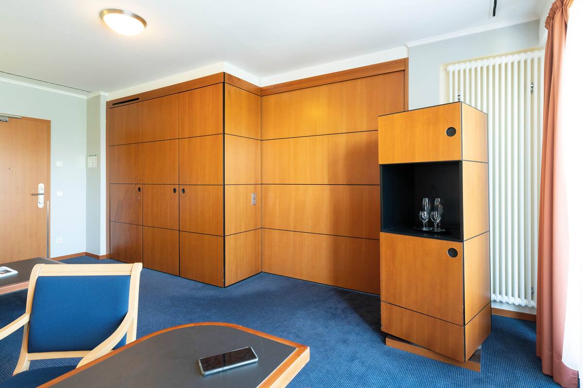 A hotel room with a large wardrobe, a small cabinet with a wine glass, a blue carpet, and a table with a phone.