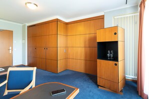 A hotel room with a large wardrobe, a small cabinet with a wine glass, a blue carpet, and a table with a phone. - Seminaris Sunday Hotel Potsdam Babelsberg (Potsdam)