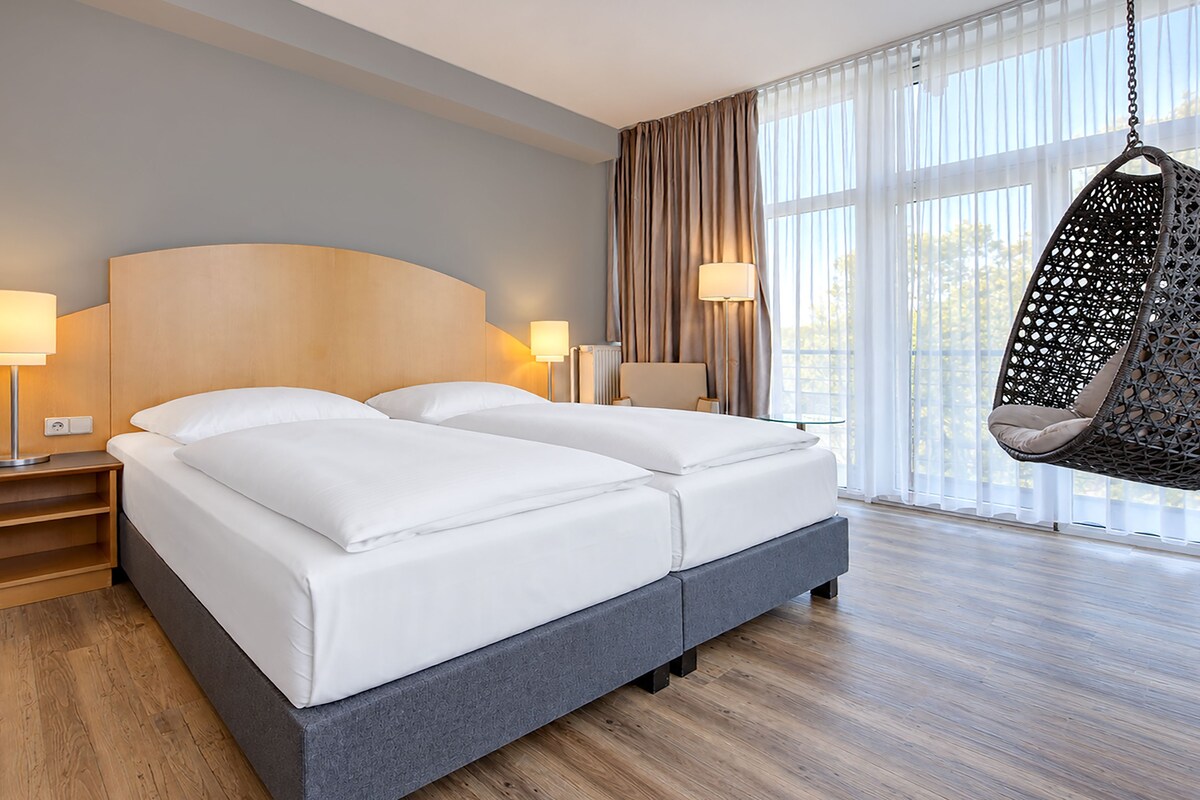 A modern hotel room with a double bed, bedside lamps, a hanging chair, and large windows.