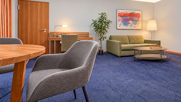 A hotel room with a dining table, two armchairs, a sofa, a small coffee table, a lamp, a plant, and a painting on the wall.