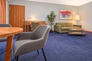 Habitación de hotel con una mesa de comedor, dos sillones, un sofá, una pequeña mesa de centro, una lámpara, una planta y un cuadro en la pared.
