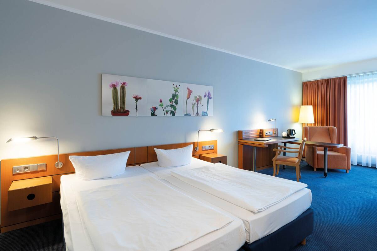 A hotel room with a double bed, a desk with a chair, a lamp, a painting on the wall, and a window with curtains.