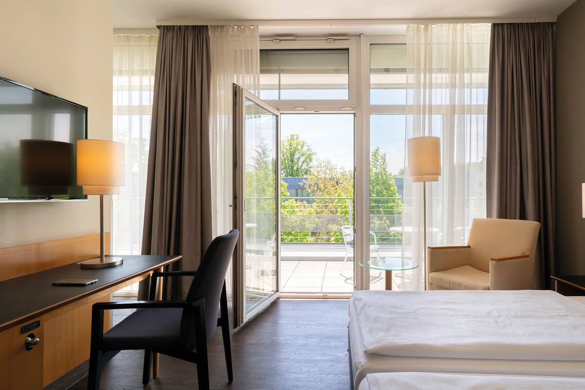 A hotel room with a bed, a desk with a chair, a television, and a view of the outdoors through large windows.