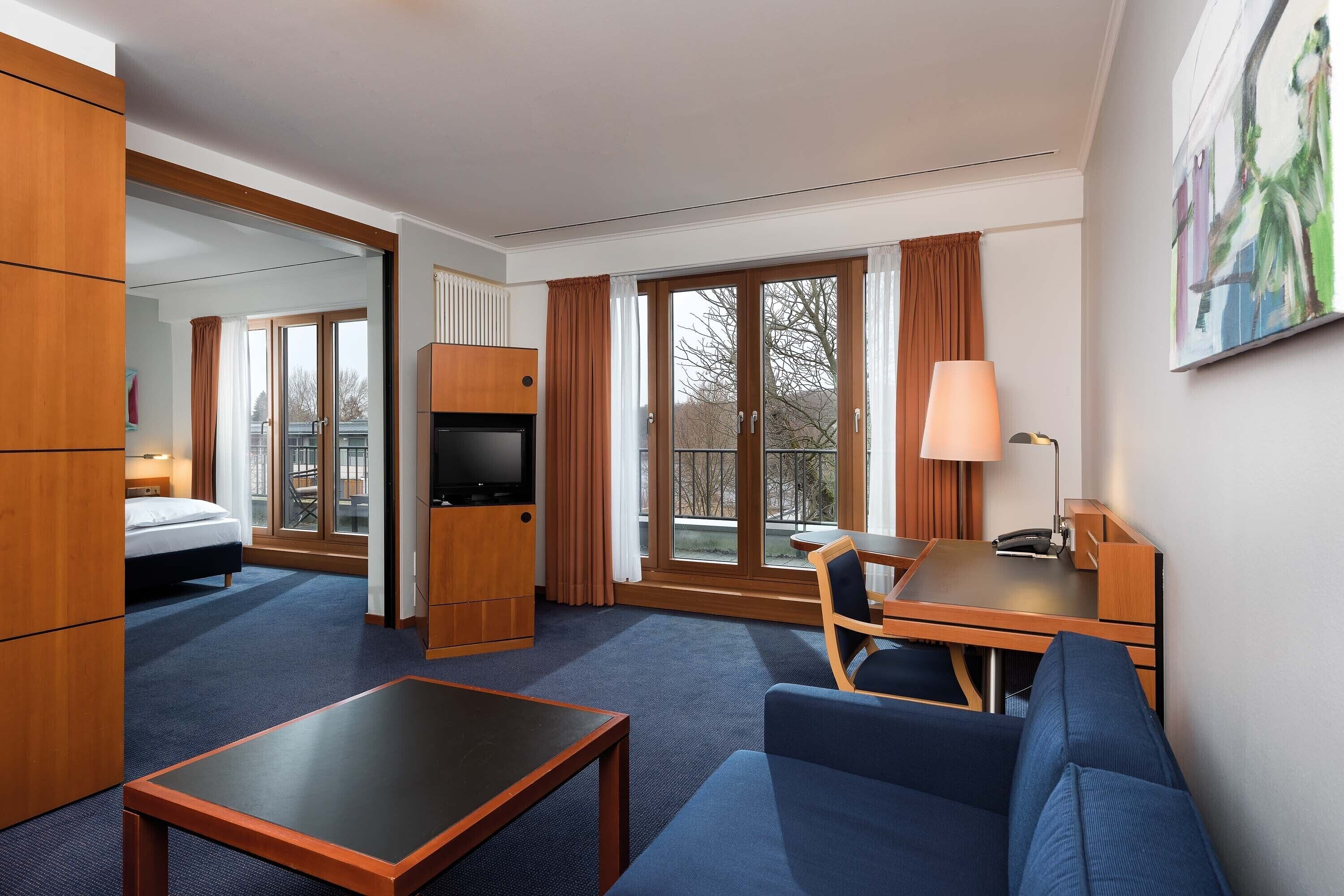 Quarto de hotel com cama, escrivaninha, sofá, televisão e janelas amplas.