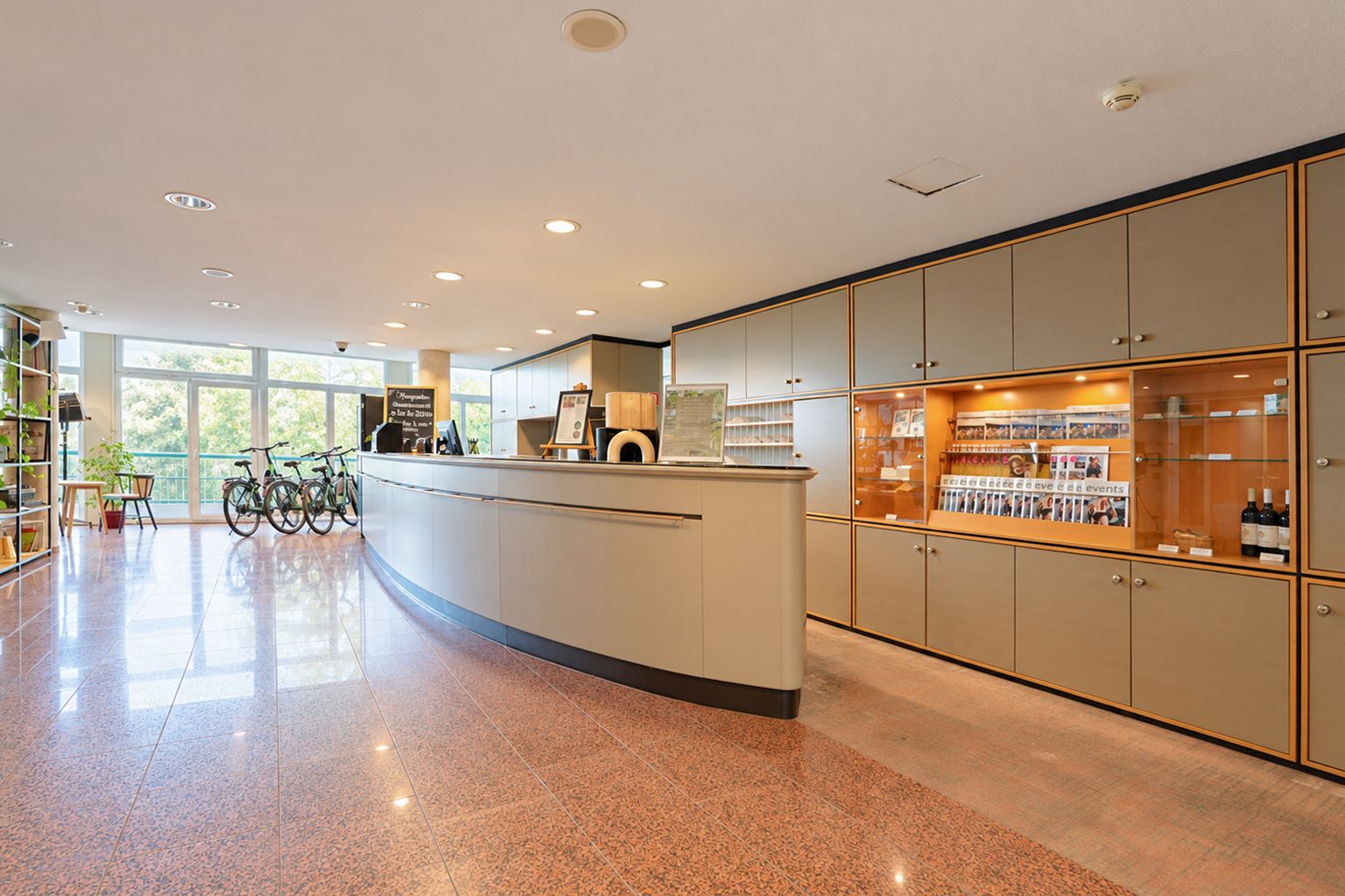 A modern hotel lobby with a curved reception desk, a glass display case with wine bottles, and bicycles parked near the entrance.