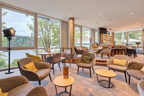 A spacious lounge area with multiple seating options, a bar, and large windows offering a view of the outdoors. - Seminaris Sunday Hotel Potsdam Babelsberg (Potsdam)