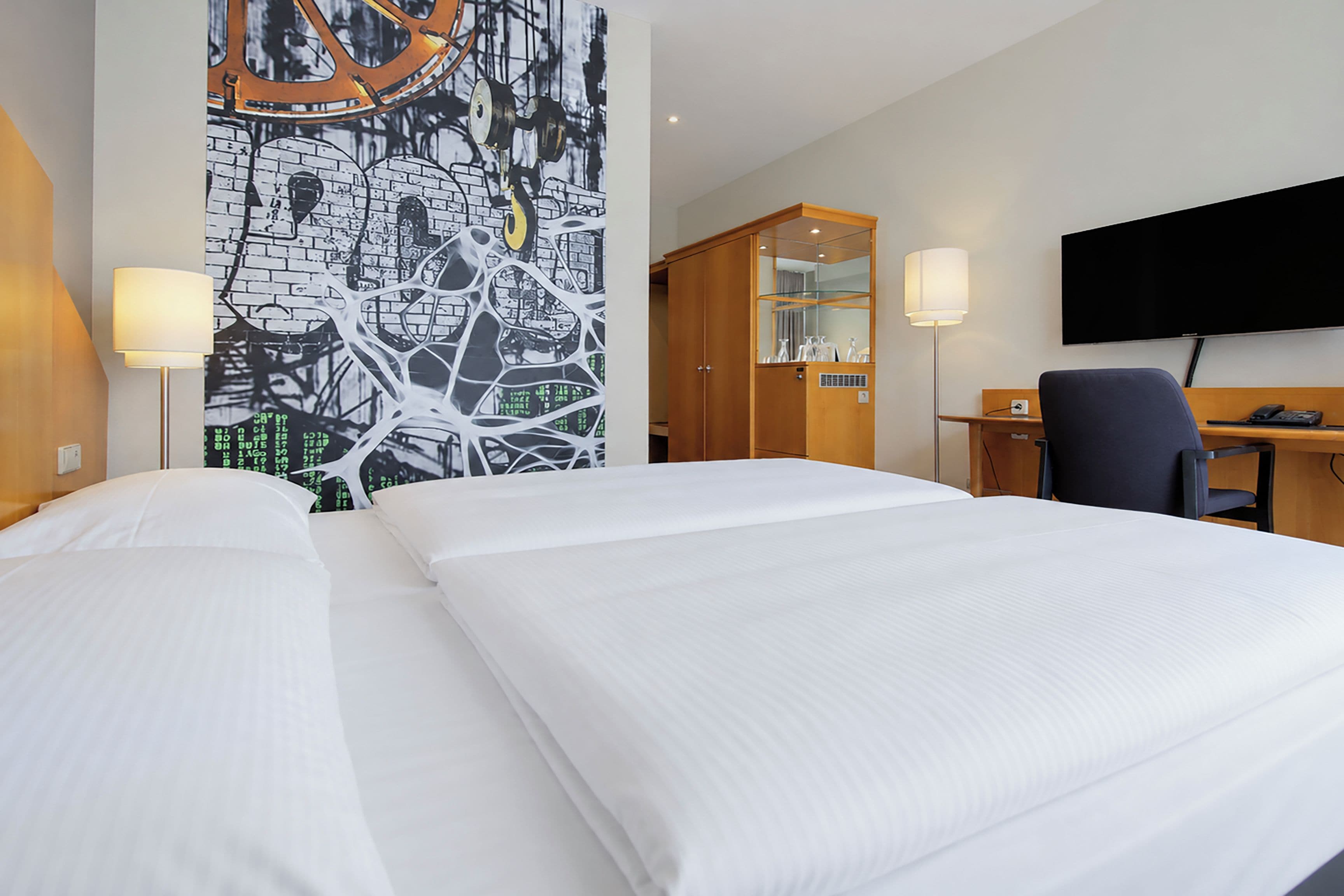 A hotel room with two beds, a desk, a chair, a TV, and a wall with a cityscape mural.