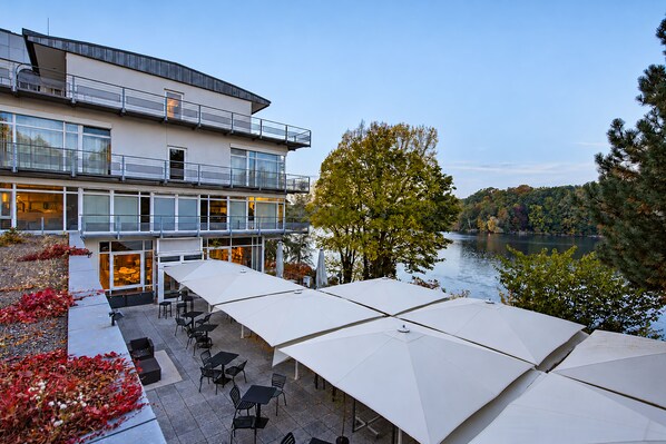 A modern building with a balcony overlooking a body of water, outdoor seating with umbrellas, and a landscaped area with trees. - Seminaris Sunday Hotel Potsdam Babelsberg (Potsdam)