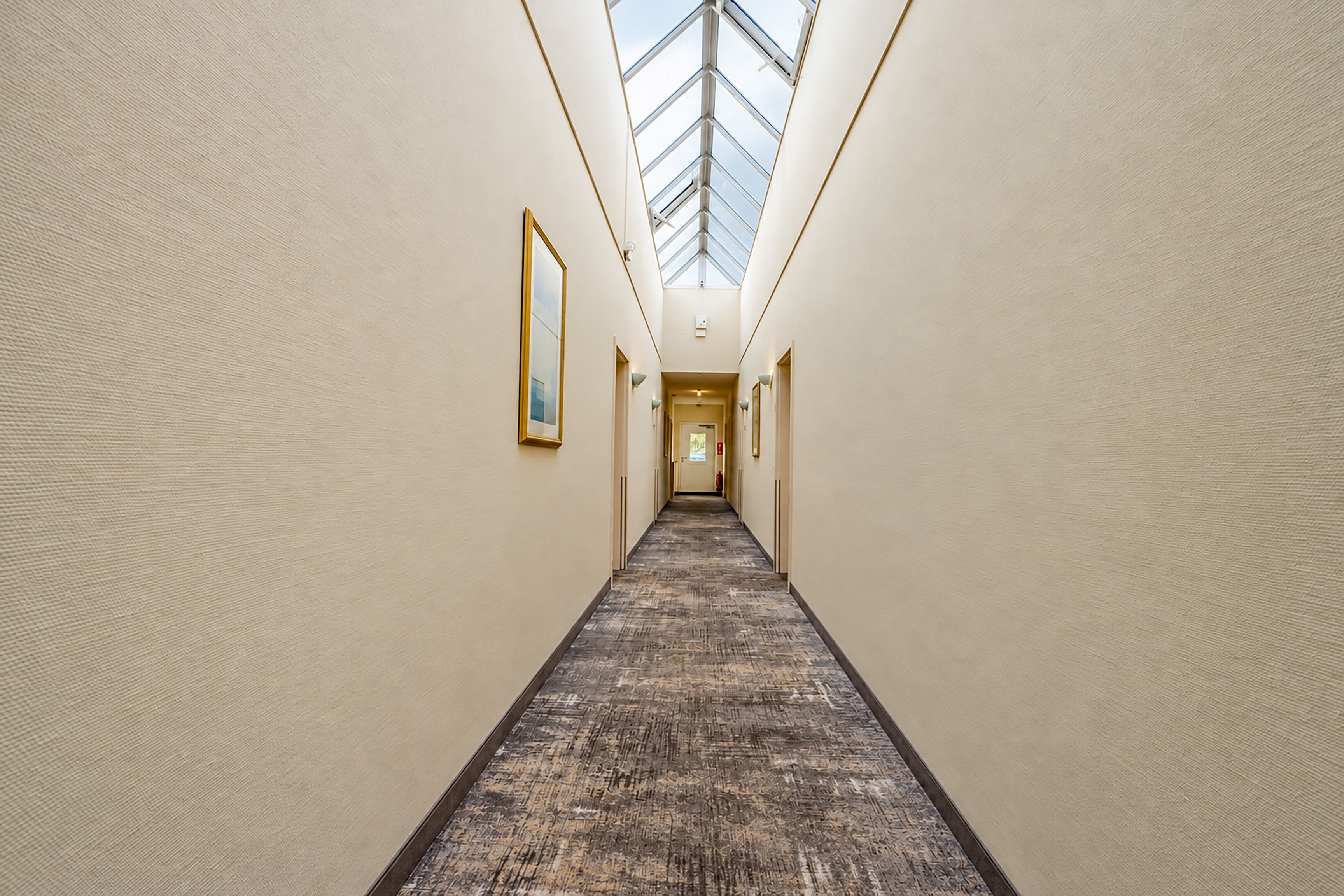 A long, narrow hallway with a patterned carpet, light-colored walls, and a skylight at the end.