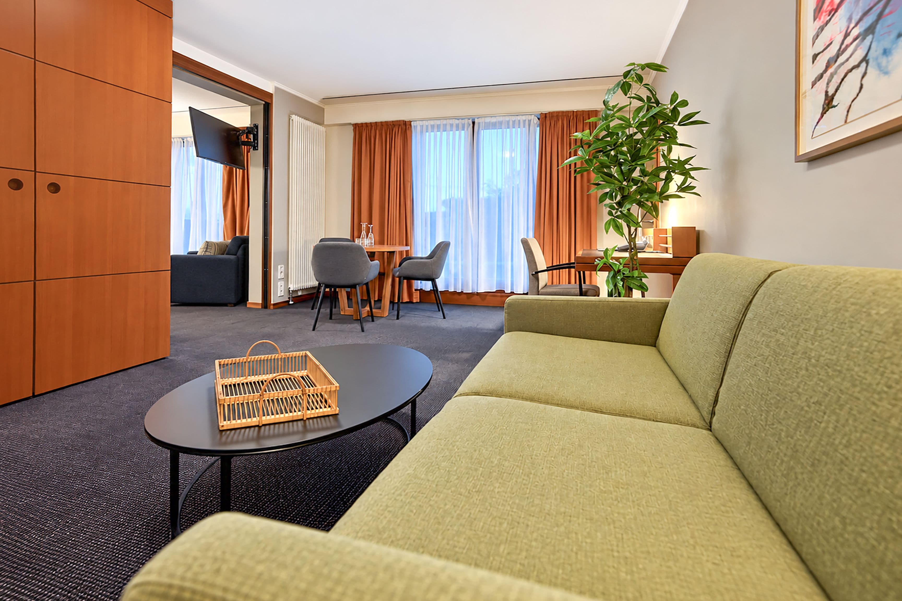 Habitación de hotel con sofá verde, una mesita redonda pequeña, una cesta de mimbre, zona de comedor con sillas, una planta y un cuadro en la pared.
