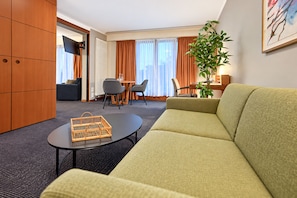 Habitación de hotel con sofá verde, una mesita redonda pequeña, una cesta de mimbre, zona de comedor con sillas, una planta y un cuadro en la pared.