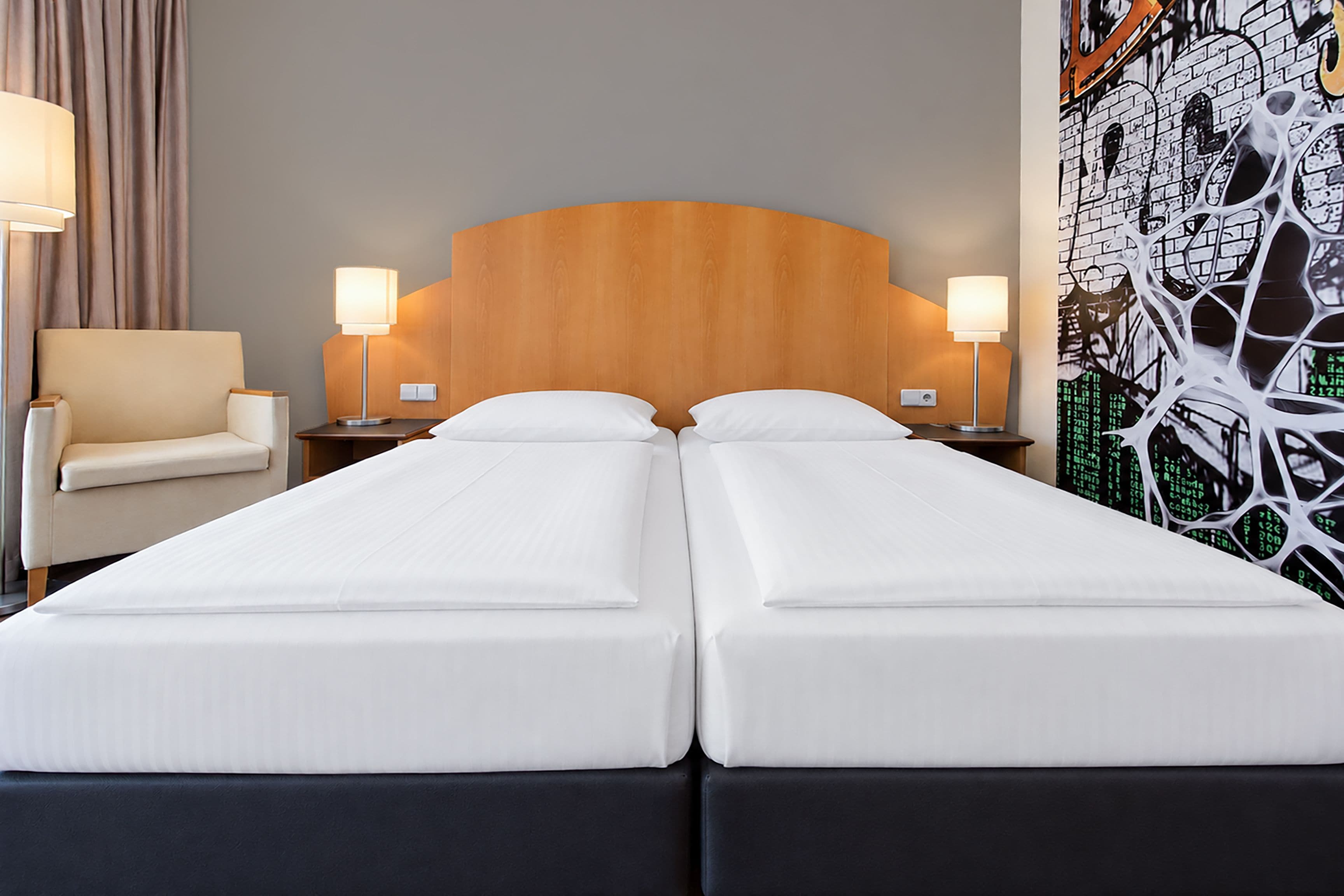 A double bed with a wooden headboard, two bedside tables with lamps, a chair, and a wall with a digital art print.