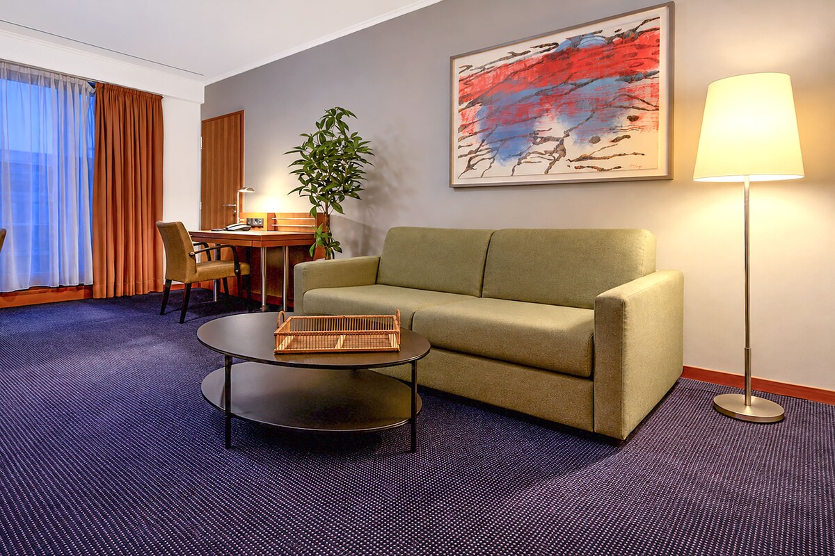 A hotel room with a sofa, a small table, a lamp, a painting, and a plant.