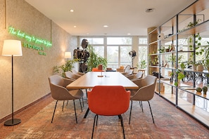 Un salón de conferencias con una larga mesa, sillas, un proyector y una estantería con plantas.