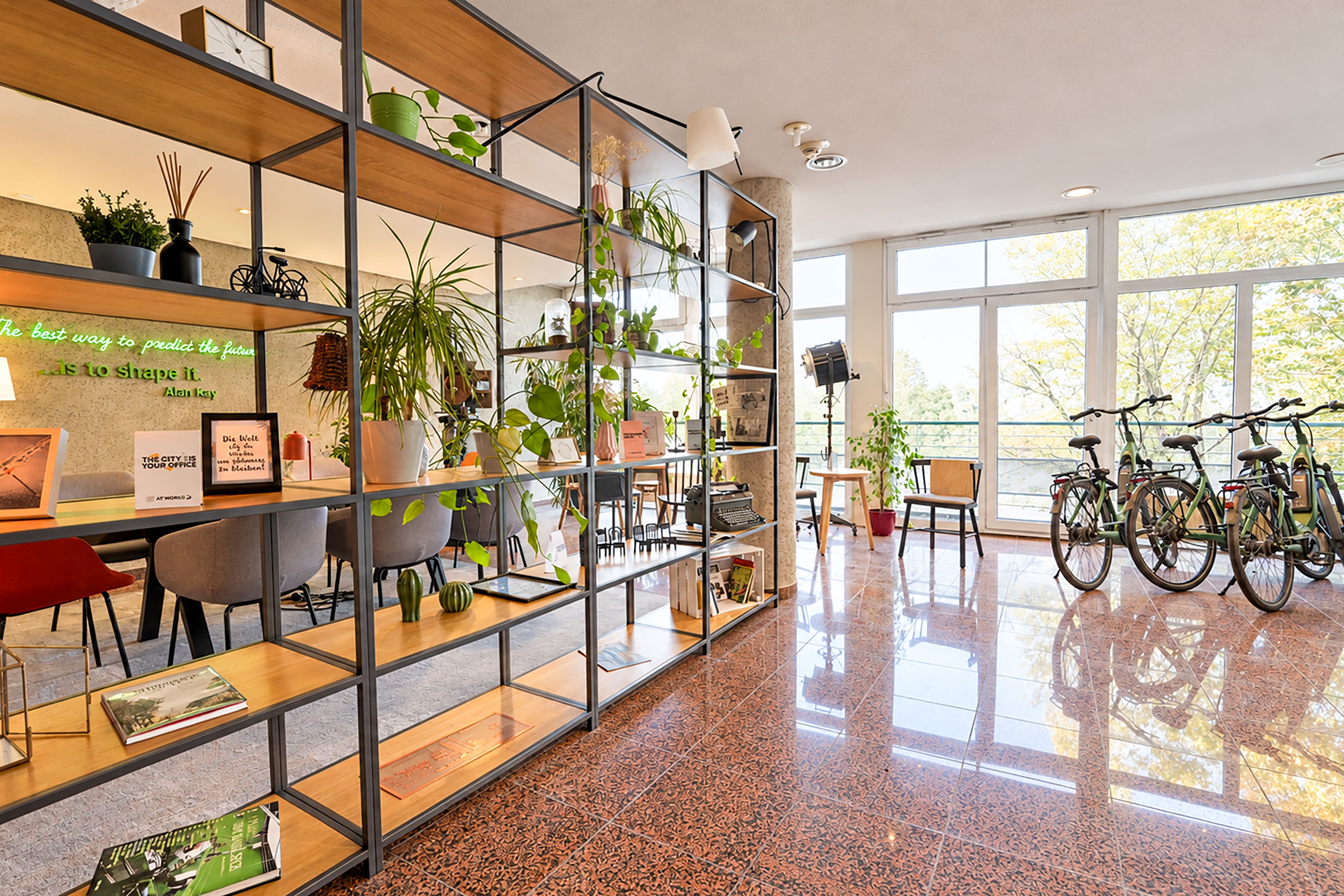 A modern office space with a glass wall, metal shelving with plants, a desk with a computer, and bicycles.