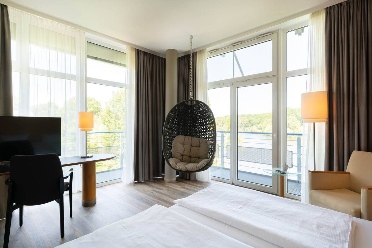 A hotel room with a bed, a desk, a chair, a hanging chair, and large windows.