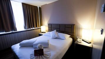 A hotel room with a neatly made bed, two bedside lamps, a telephone, and a window with curtains.