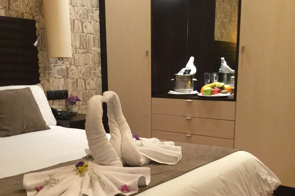 Een hotelkamer met een netjes opgemaakt bed, een houten nachtkastje met een lamp, een minibar en uitzicht op een keuken met fruit en een pan.