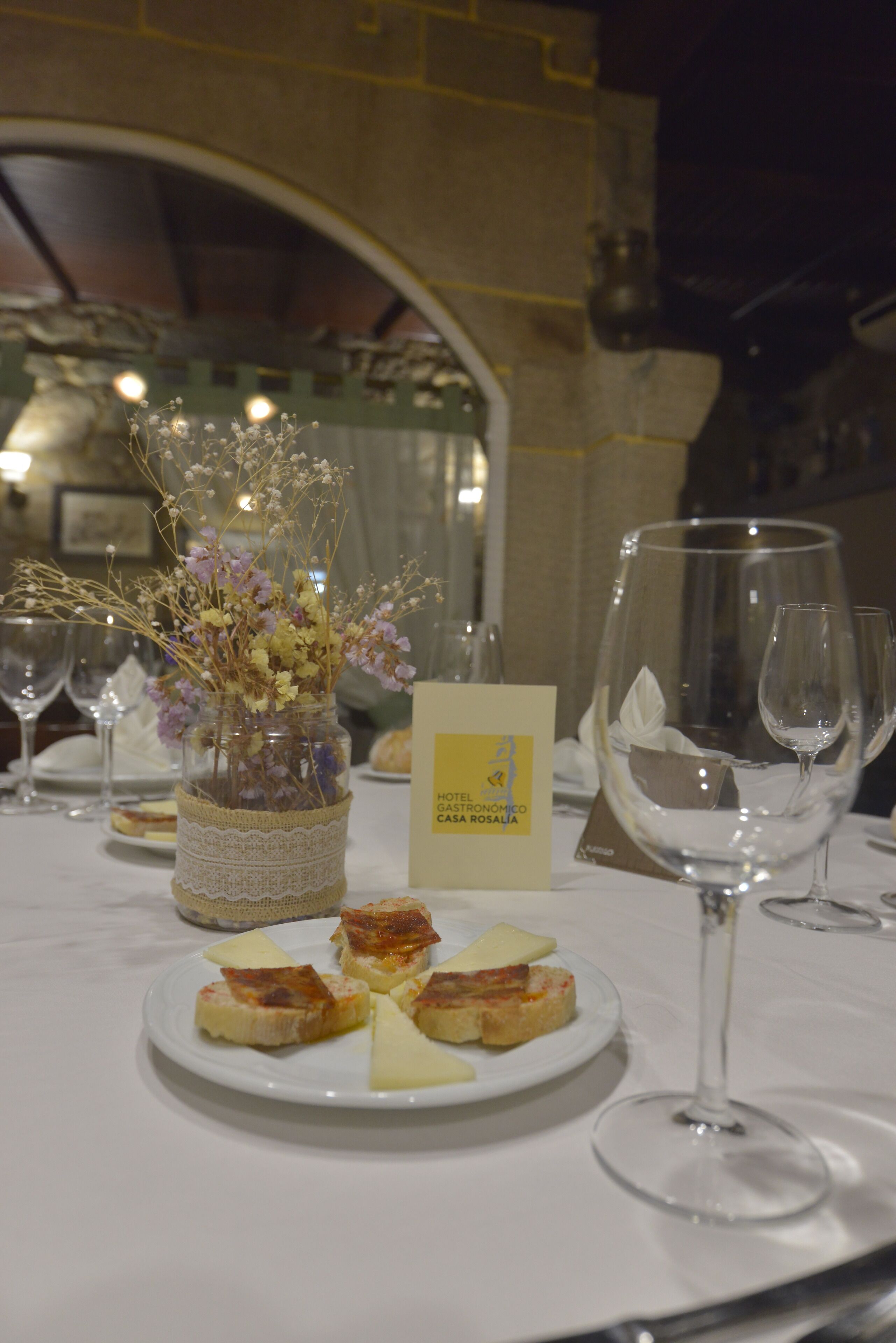 A table set with a small vase of dried flowers, a plate of cheese, and wine glasses.