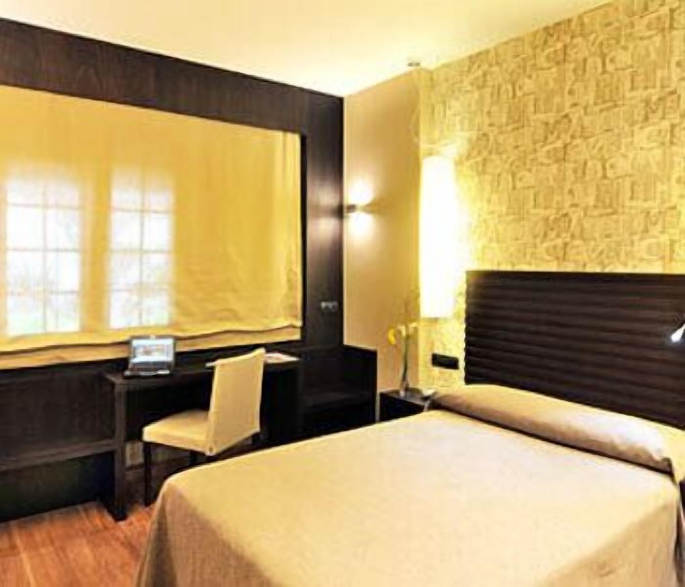 A hotel room with a bed, a desk, a chair, a lamp, and a window with curtains.