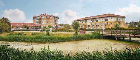 A residential building with multiple floors and balconies, a bridge, and a grassy area with water.