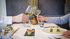 A dining table with a white tablecloth, two people toasting with wine glasses, and a variety of dishes including a bowl of nuts and a plate with a green dessert. - Greenside (De Koog)