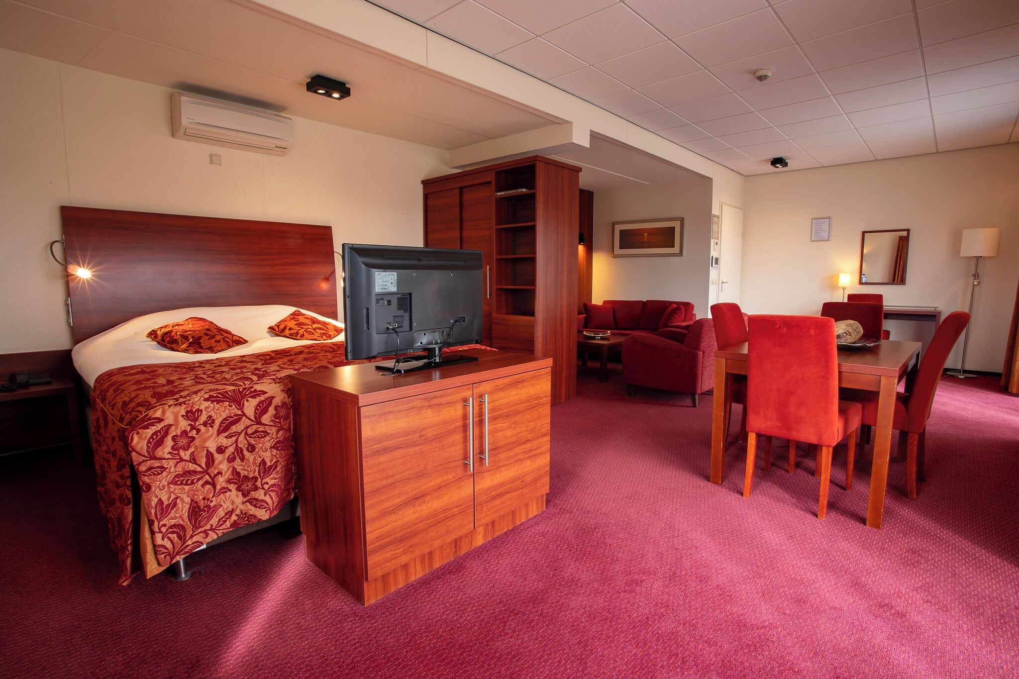 Een hotelkamer met een bed, een televisie op een houten dressoir, een rode bank, een eettafel met stoelen en een klein bureau met een lamp.