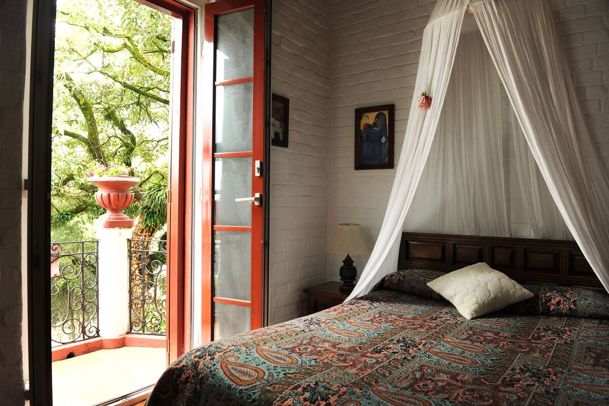 A bedroom with a bed, a nightstand, a lamp, a window with a view of trees, and a door.