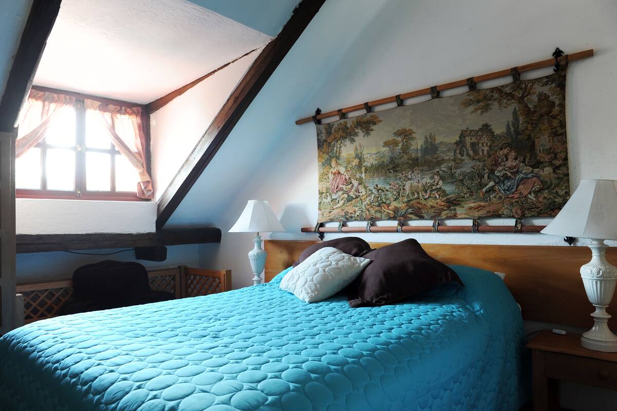 A bedroom with a blue quilted bedspread, a wooden headboard, a painting above the bed, and a window with curtains.