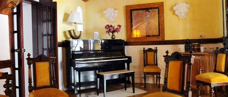 A room with a piano, wooden chairs, a bench, a table with a magazine, and a wall with decorative elements.