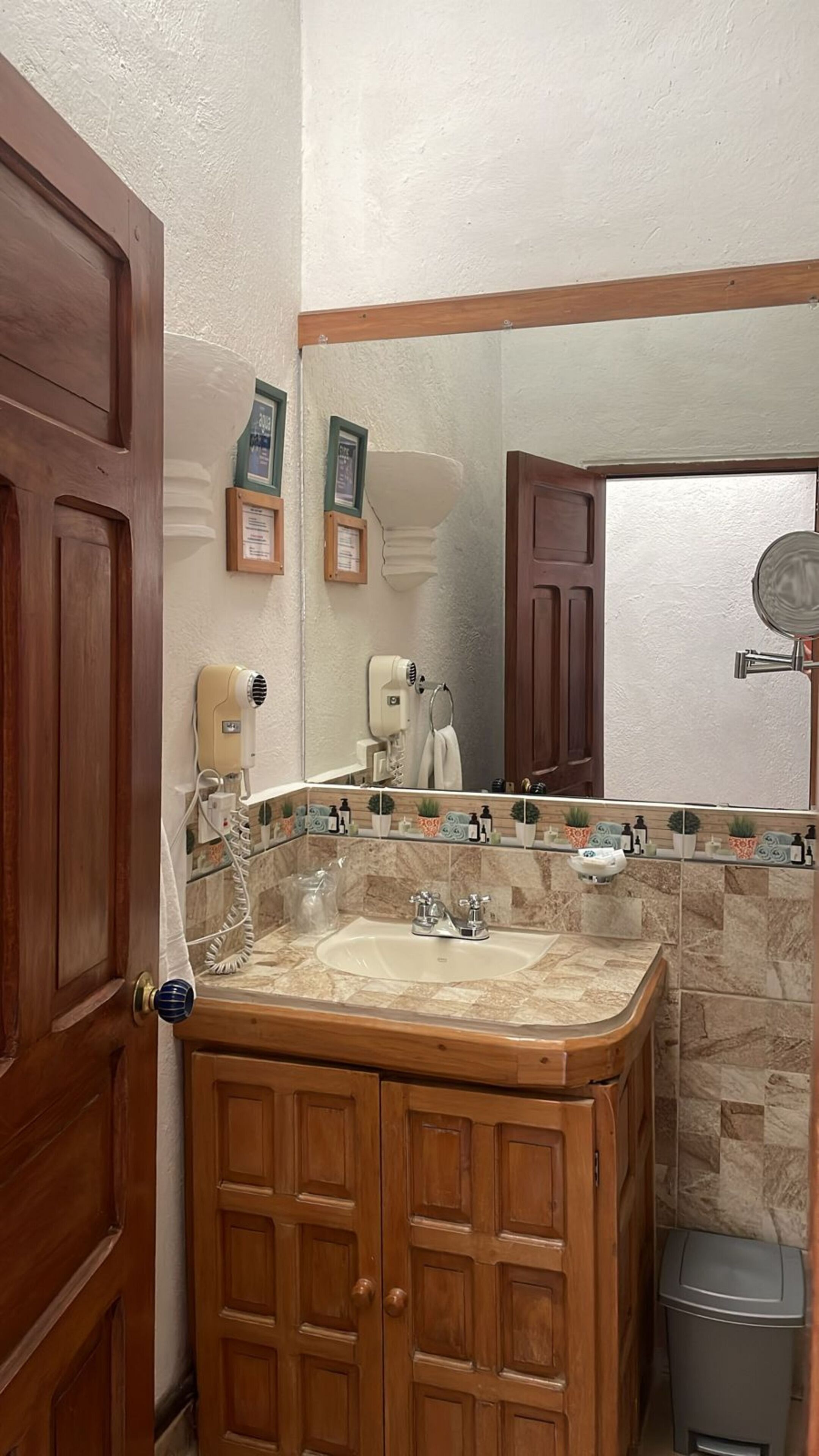 A bathroom with a wooden vanity, a sink, and a mirror. There are framed certificates on the wall, a hairdryer, and a trash bin.