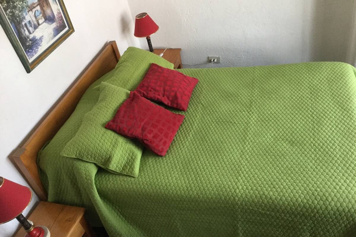 A bed with a green quilt and two red pillows, a wooden nightstand with a red lamp, and a painting on the wall.