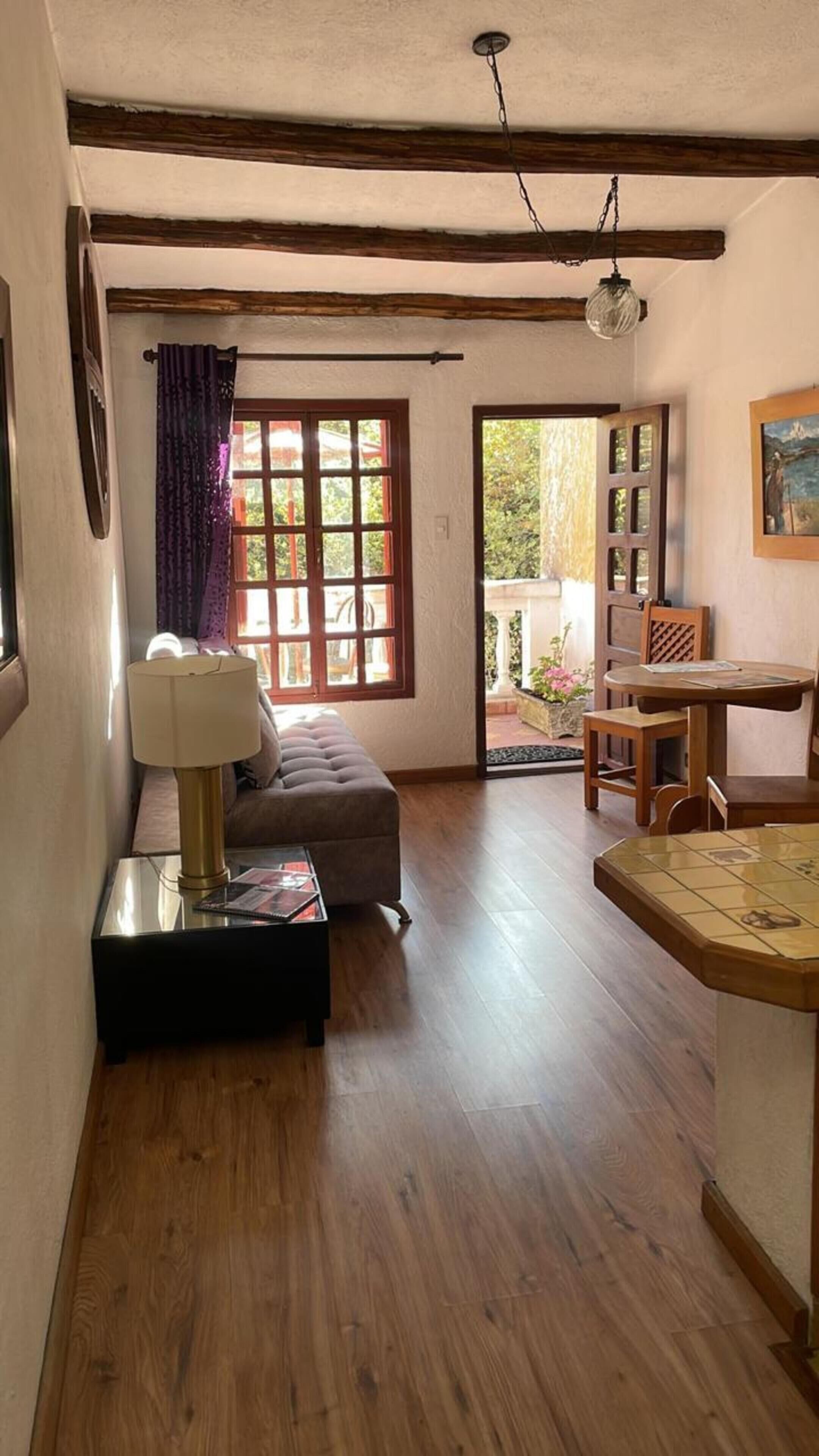 A cozy living room with a sofa, a table, and a lamp. There is a window with curtains and a view of the outdoors.