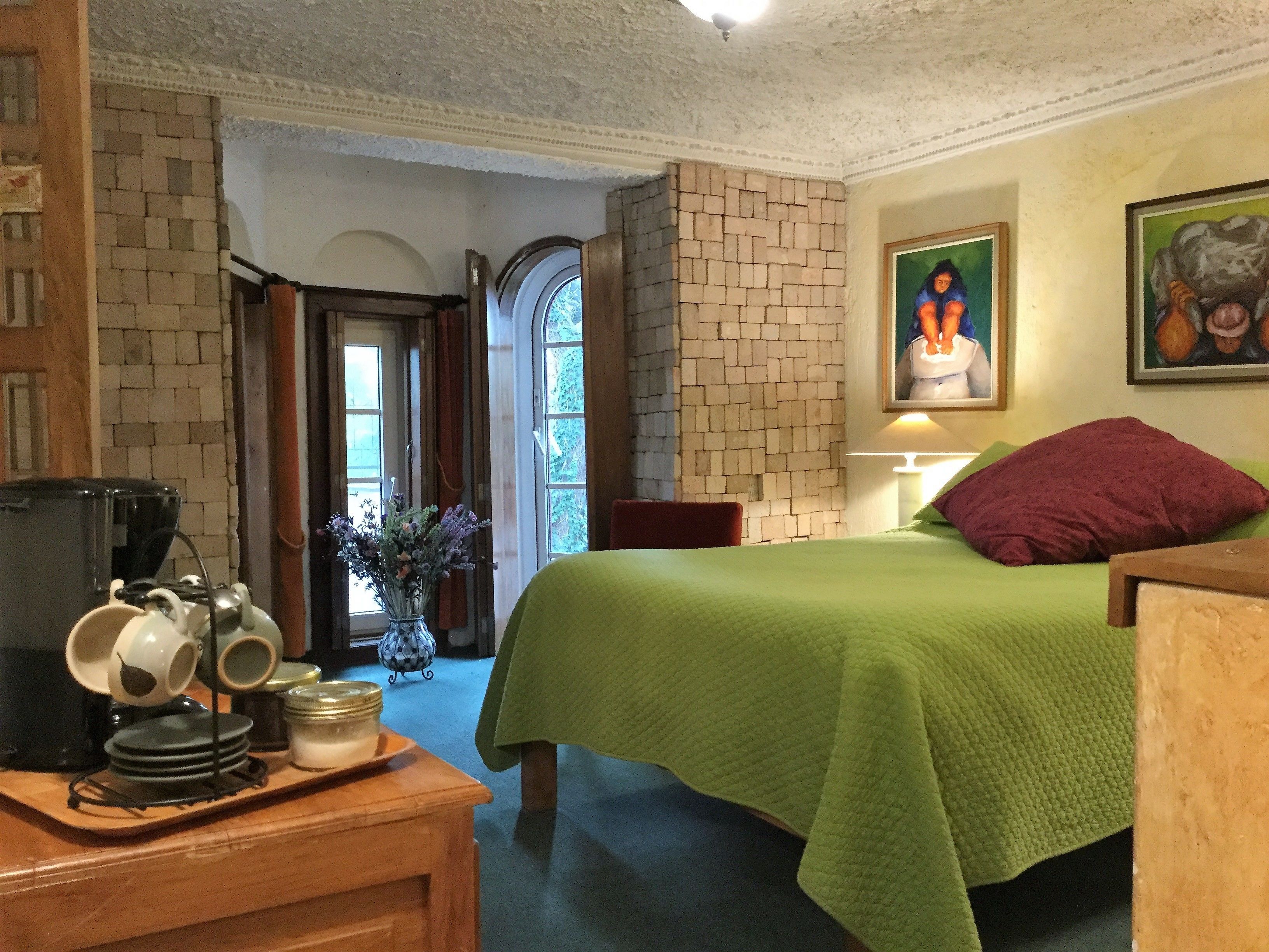 A hotel room with a green bedspread, a wooden nightstand with a coffee maker, a vase with flowers, and a painting on the wall.