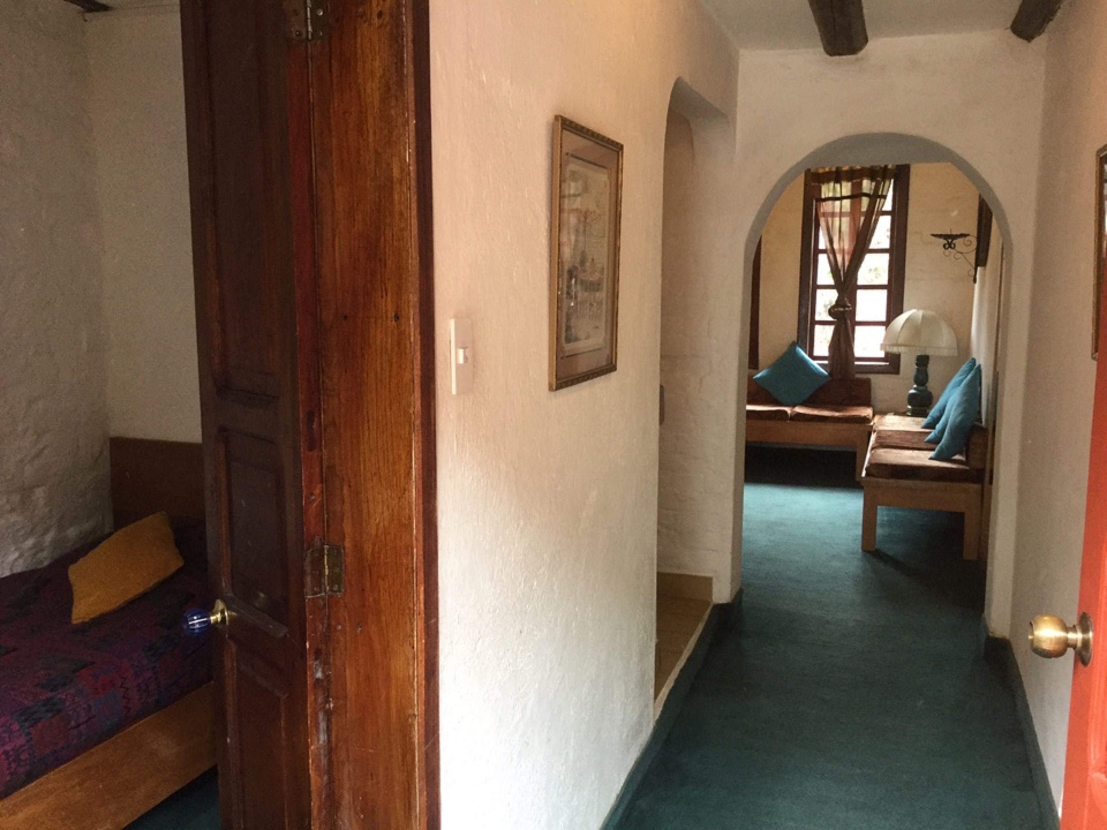 Un couloir avec une porte en bois, un tableau encadré au mur, une moquette bleue et un coin salon avec une table et des chaises.