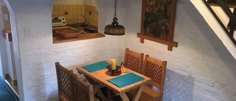 A dining area with a table and chairs, a hanging light fixture, and a kitchen visible through an archway.