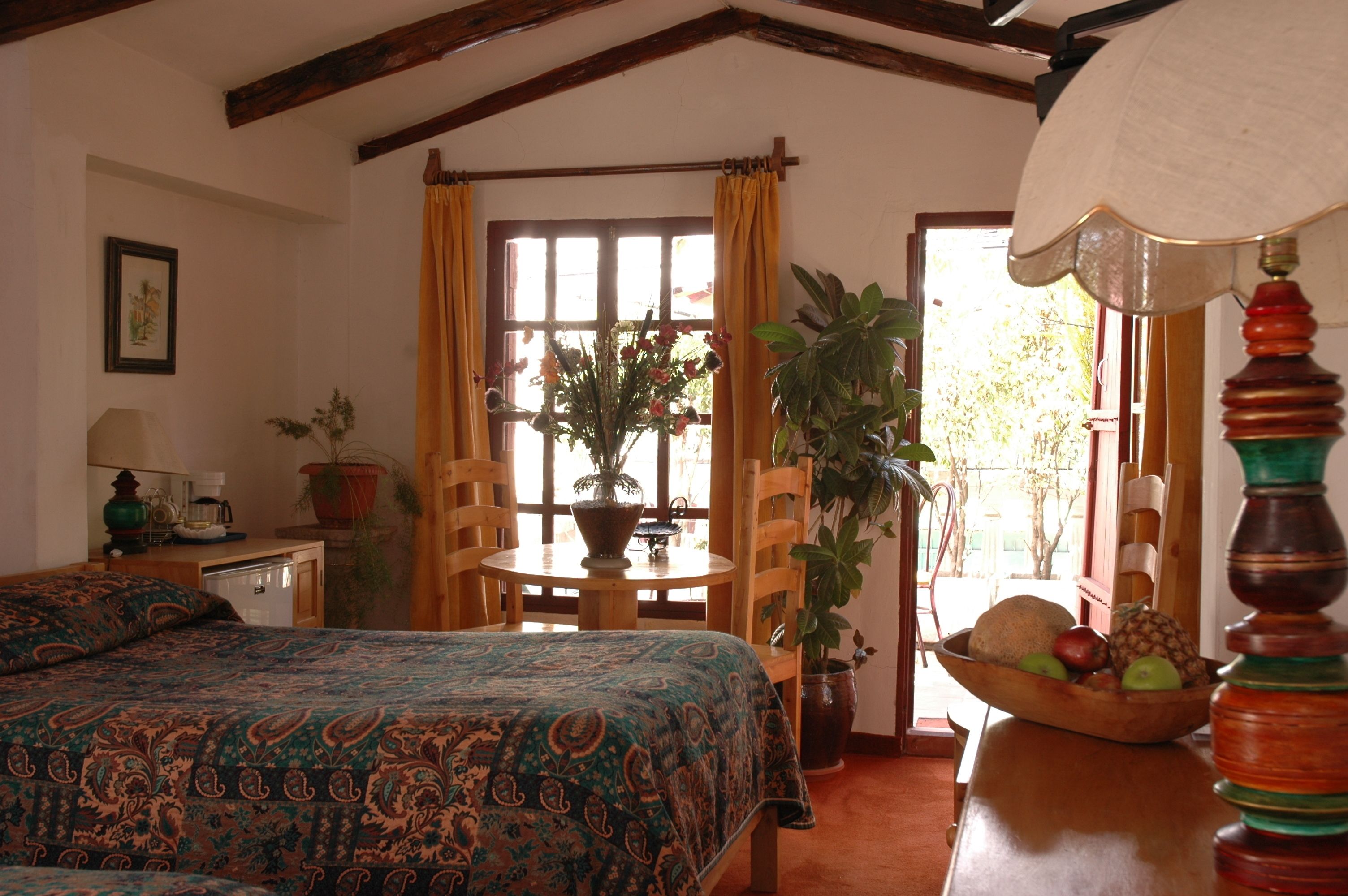 Una habitación con una cama, una mesa con frutero, una planta en maceta y una lámpara.
