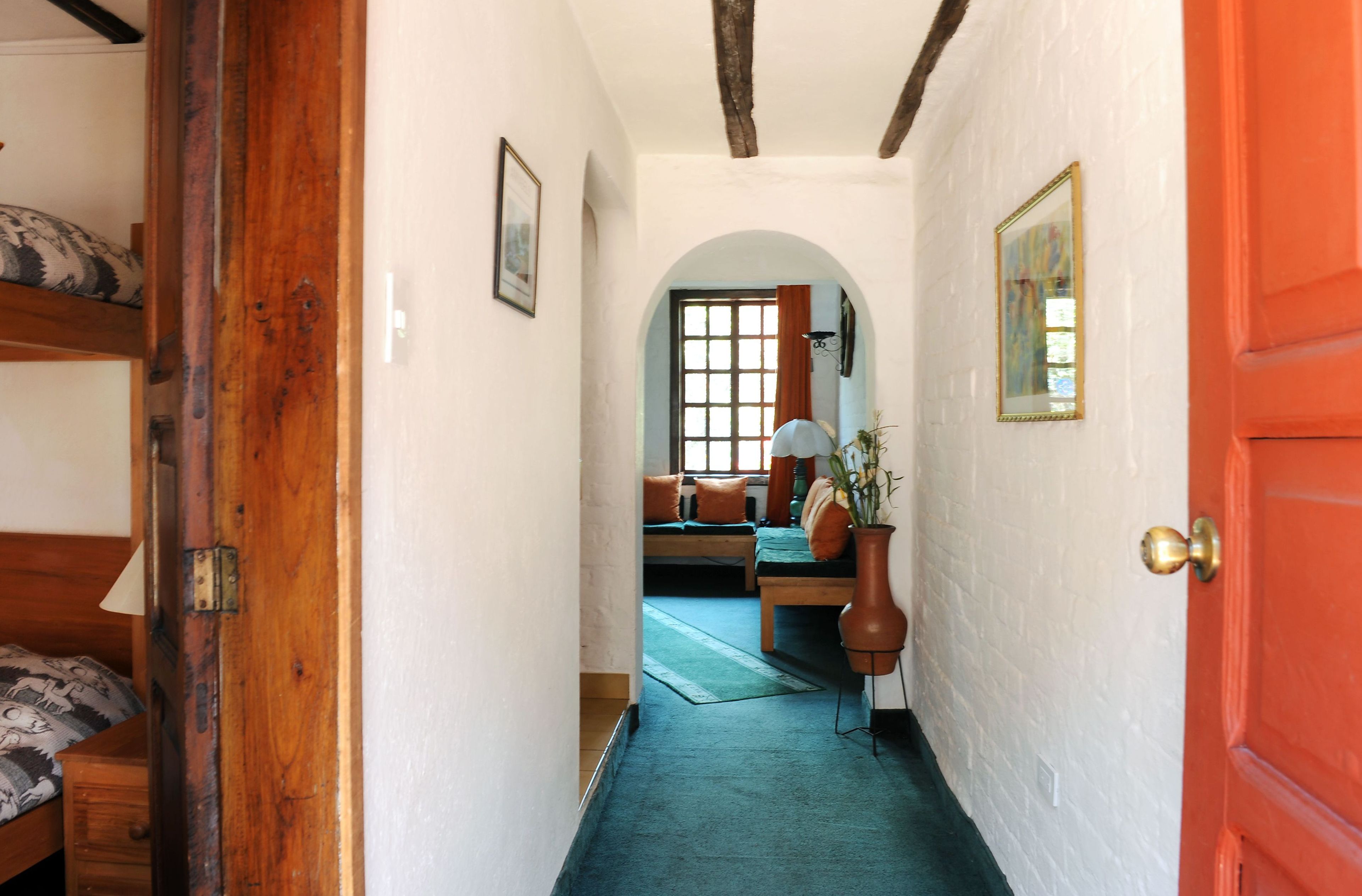 A narrow hallway with a wooden door on the left, a red door on the right, and a bed with a patterned bedspread at the end.