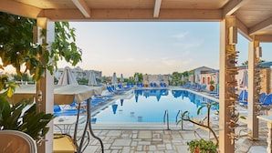 A pool area with lounge chairs, a covered seating area, and a view of buildings in the background. - Golden Bay Apartments-Garden Haven (Hersonissos)