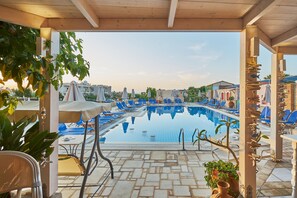 A pool area with lounge chairs, a covered seating area, and a view of buildings in the background. - Golden Bay Apartments-Garden Haven (Hersonissos)
