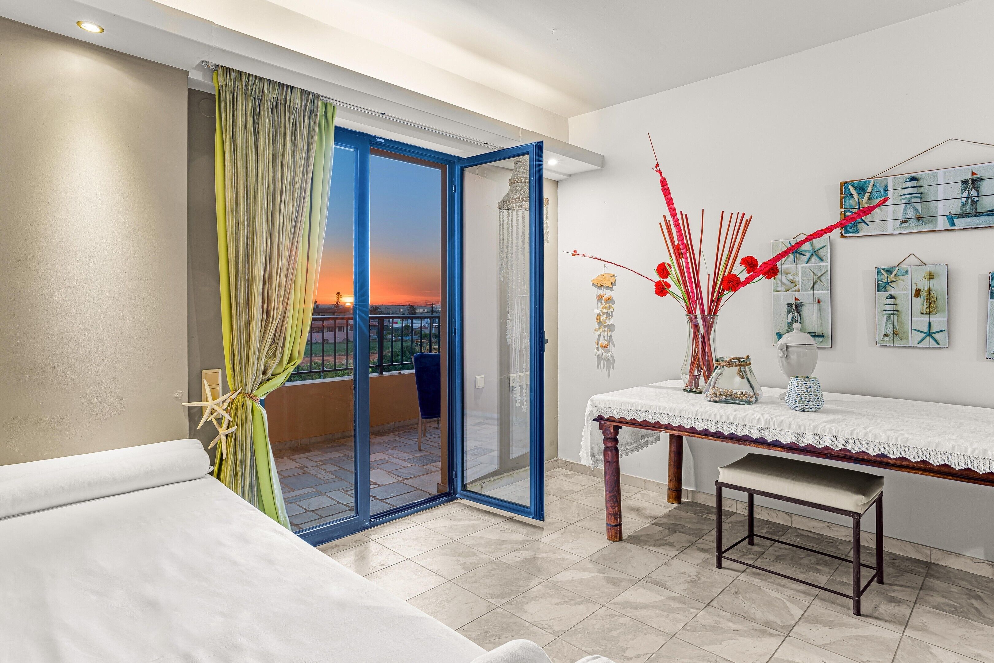 A balcony with a view of the sunset, a sliding glass door, a white table with a bench, and decorative wall art.