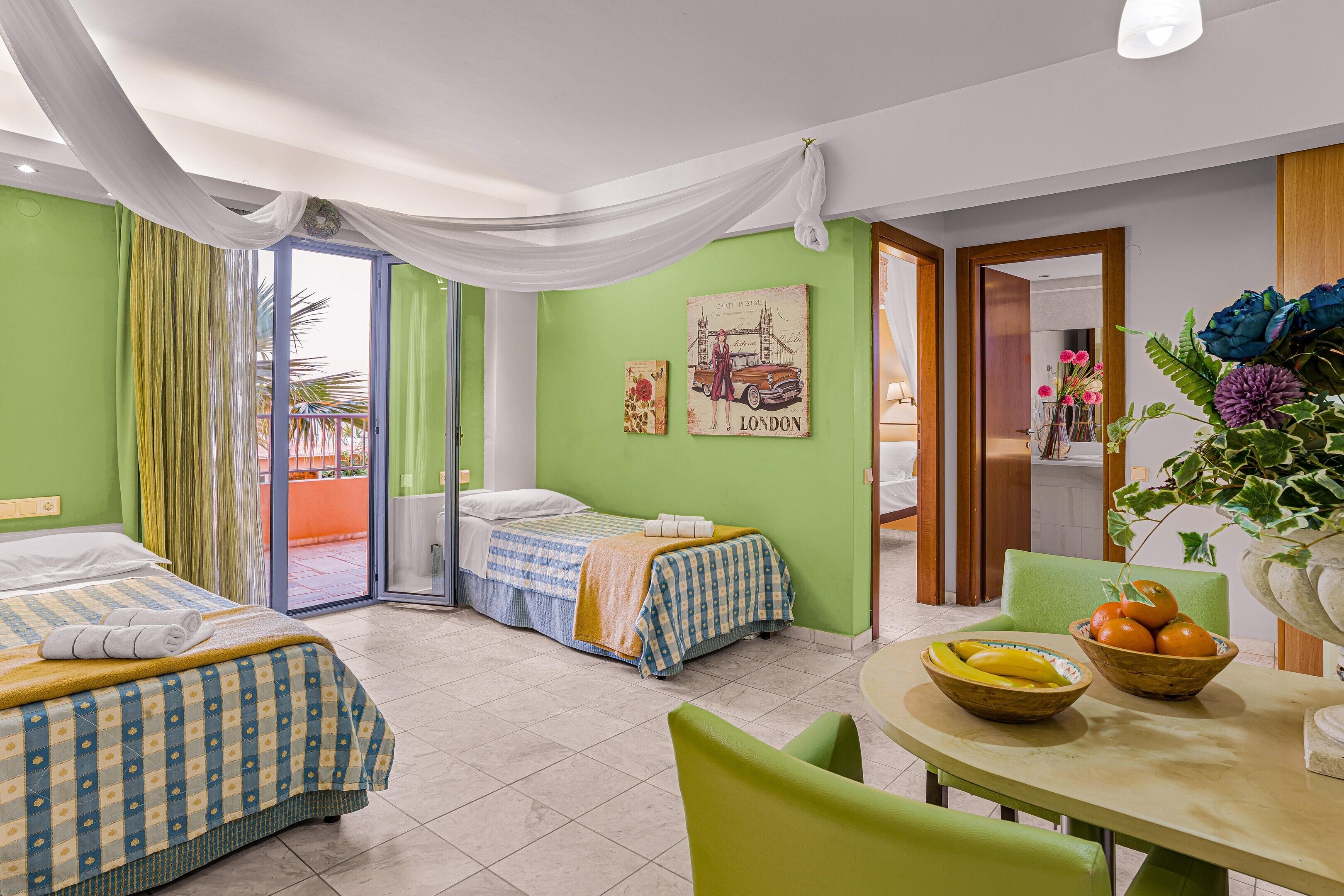 A hotel room with two beds, a dining table with fruit, and a view of the outdoors.