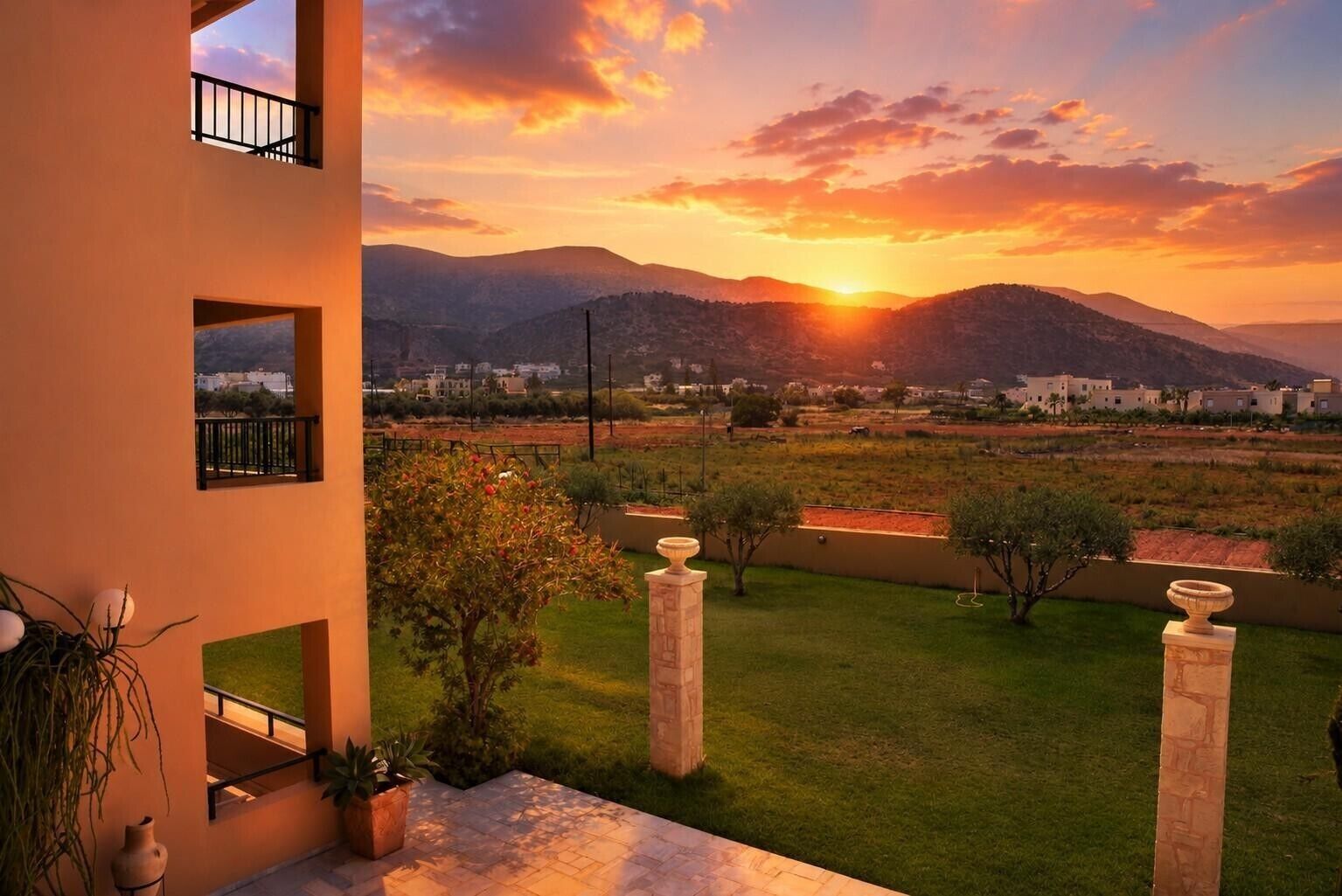 A balcony with a view of a sunset over a hilly landscape, a grassy area, and a building in the distance.