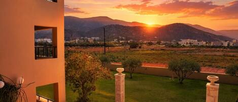 A balcony with a view of a sunset over a hilly landscape, a grassy area, and a building in the distance.