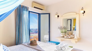 A hotel room with a bed, a window with curtains, a mirror, and a small table with a plant and decorative items. - Golden Bay Apartments-Garden Haven (Hersonissos)