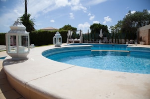 A swimming pool with a curved edge, surrounded by a white deck. There are two white lanterns and a canopy structure in the background.
