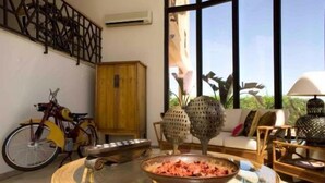 A room with a large window, a wooden cabinet, a bicycle, a round table with a bowl, and a sofa with cushions. - Parque Das Laranjeiras (Loulé)