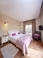 Un dormitorio con cama, mesitas de noche, una silla, televisor y ventana con cortinas.