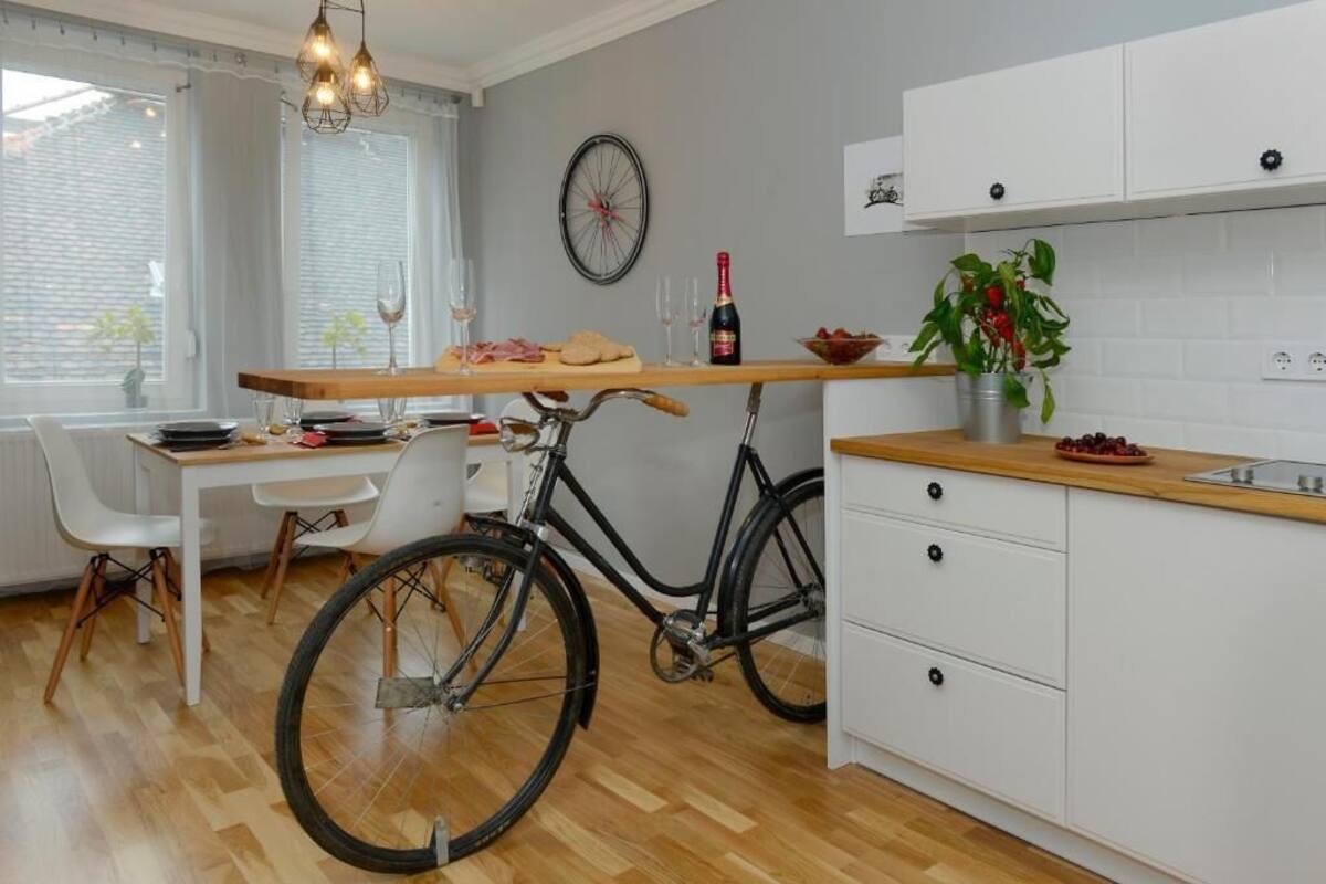 A modern kitchen with a bicycle, white cabinets, a wooden dining table, and a window with blinds.