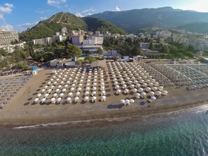 Une vue aérienne d’une plage, avec des rangées de parasols et de chaises longues, une mer bleue et limpide, et des bâtiments en arrière-plan.